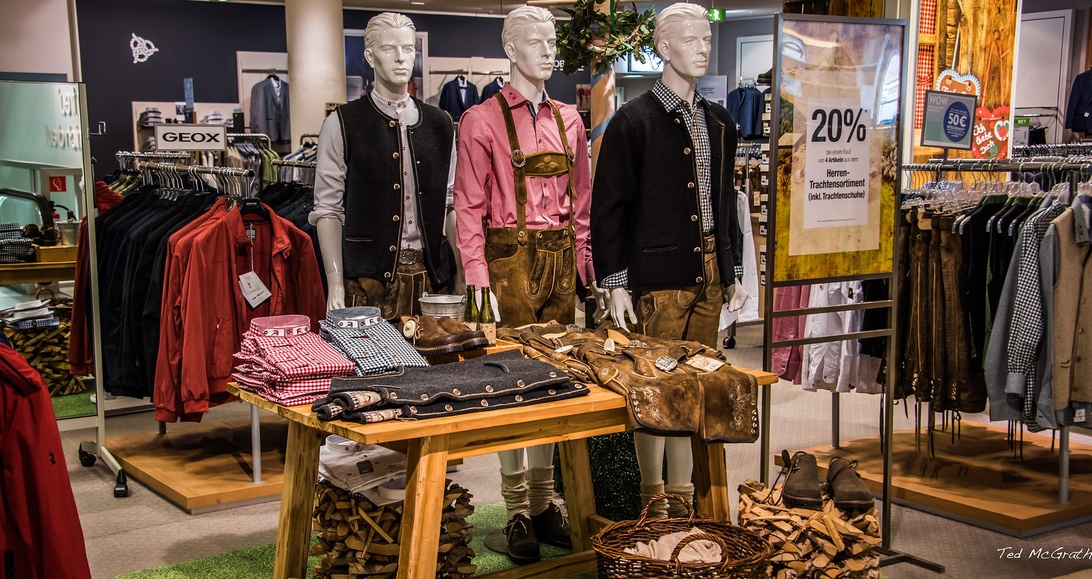 Butiksfönster med skyltdockor klädda i traditionella tyska dräkter, inklusive bruna lederhosen och rutiga skjortor.